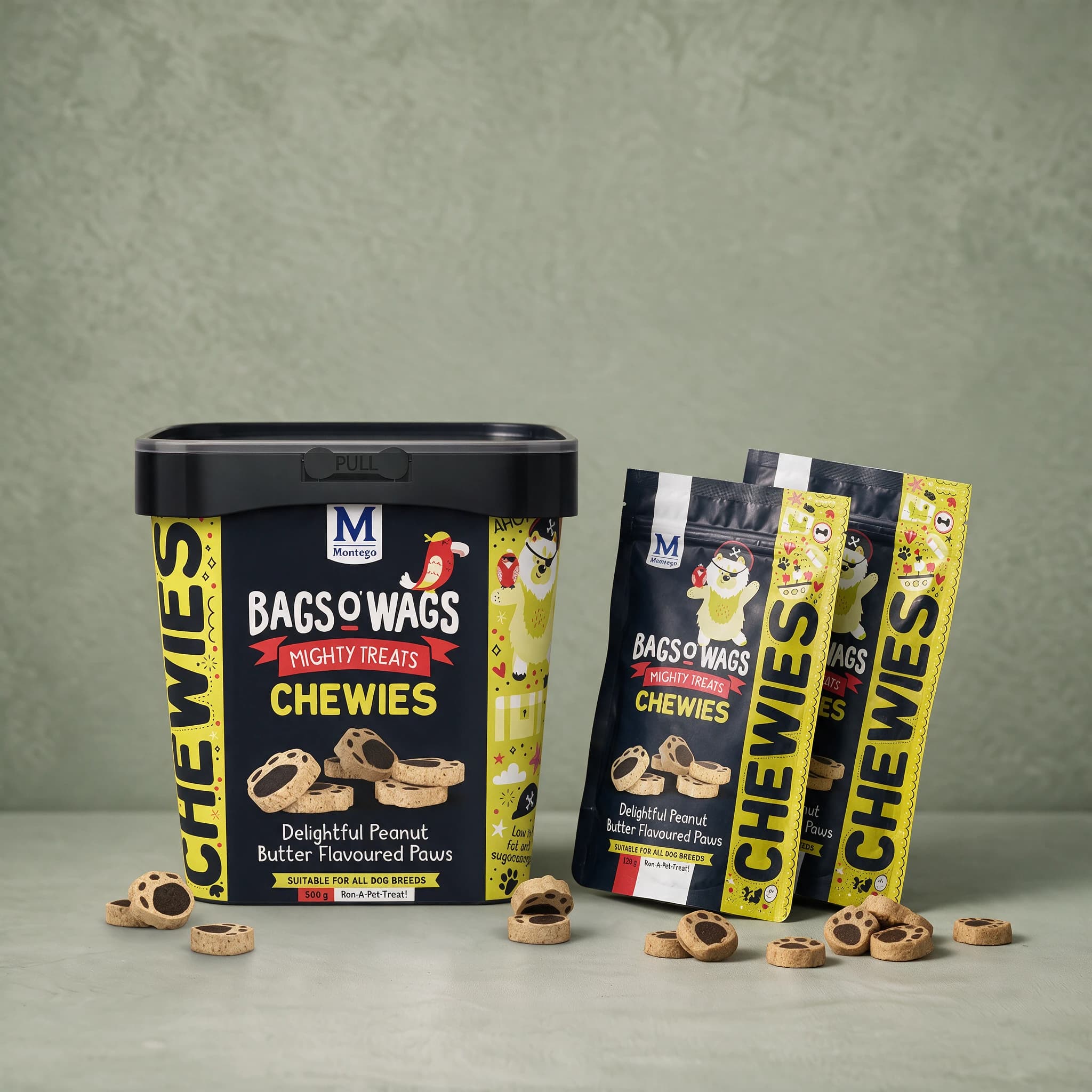 A black plastic 500 g tub of Bags O' Wags Mighty Treats Chewies Peanut Butter Paws standing beside two black 120 g pouches of the same flavour, all three packs against a plain sage green studio background.