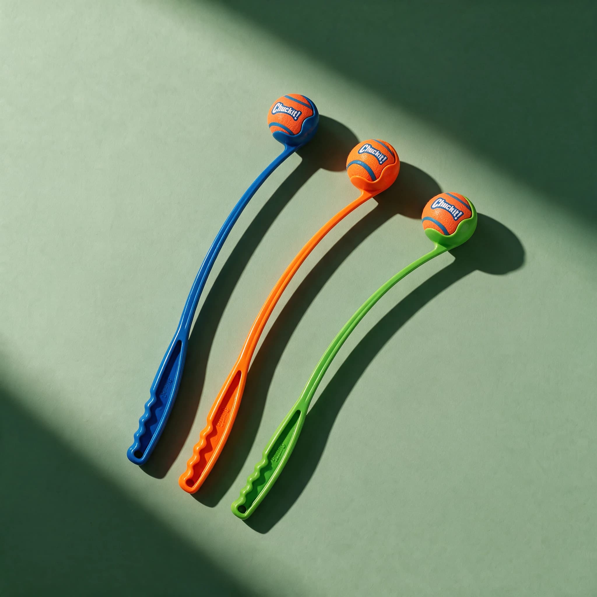 Three Chuckit Classic Launchers laid out together in blue, orange, and green, each with a matching orange and blue Chuckit tennis ball cupped in the curved scoop end.