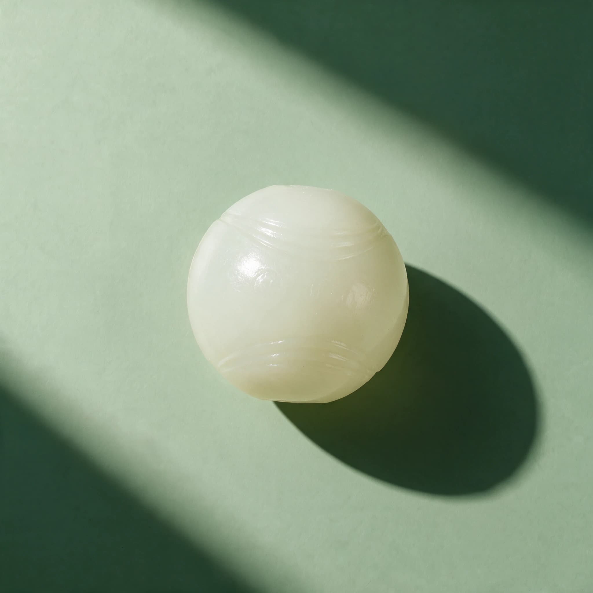 Overhead view of a single cream-white Chuckit! Max Glow Ball with a ridged surface texture, resting on a sage-green background with a dramatic diagonal shadow.