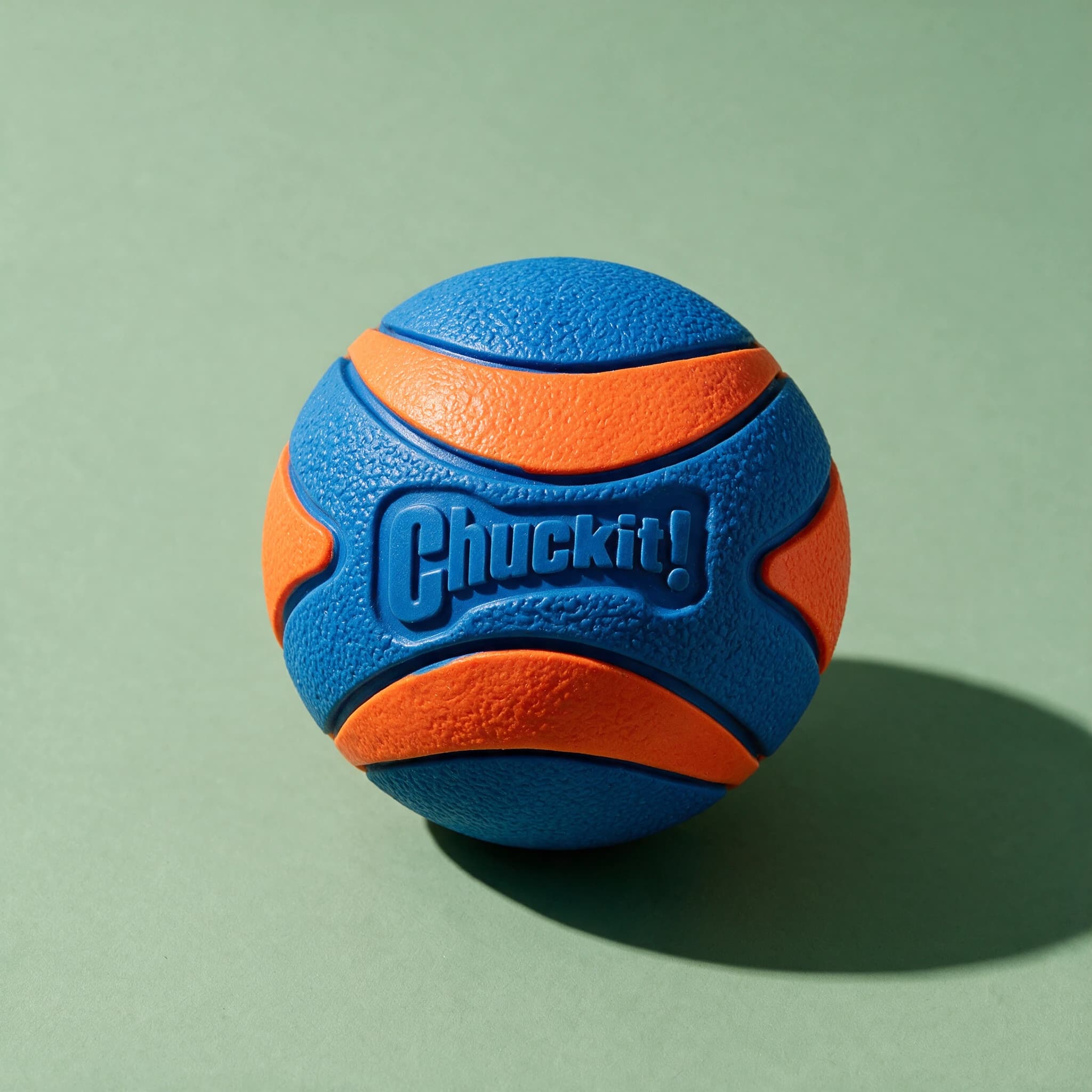 Close-up studio shot of the blue and orange textured Chuckit! Ultra Squeaker Ball with the embossed "Chuckit!" logo, set against a sage green background.