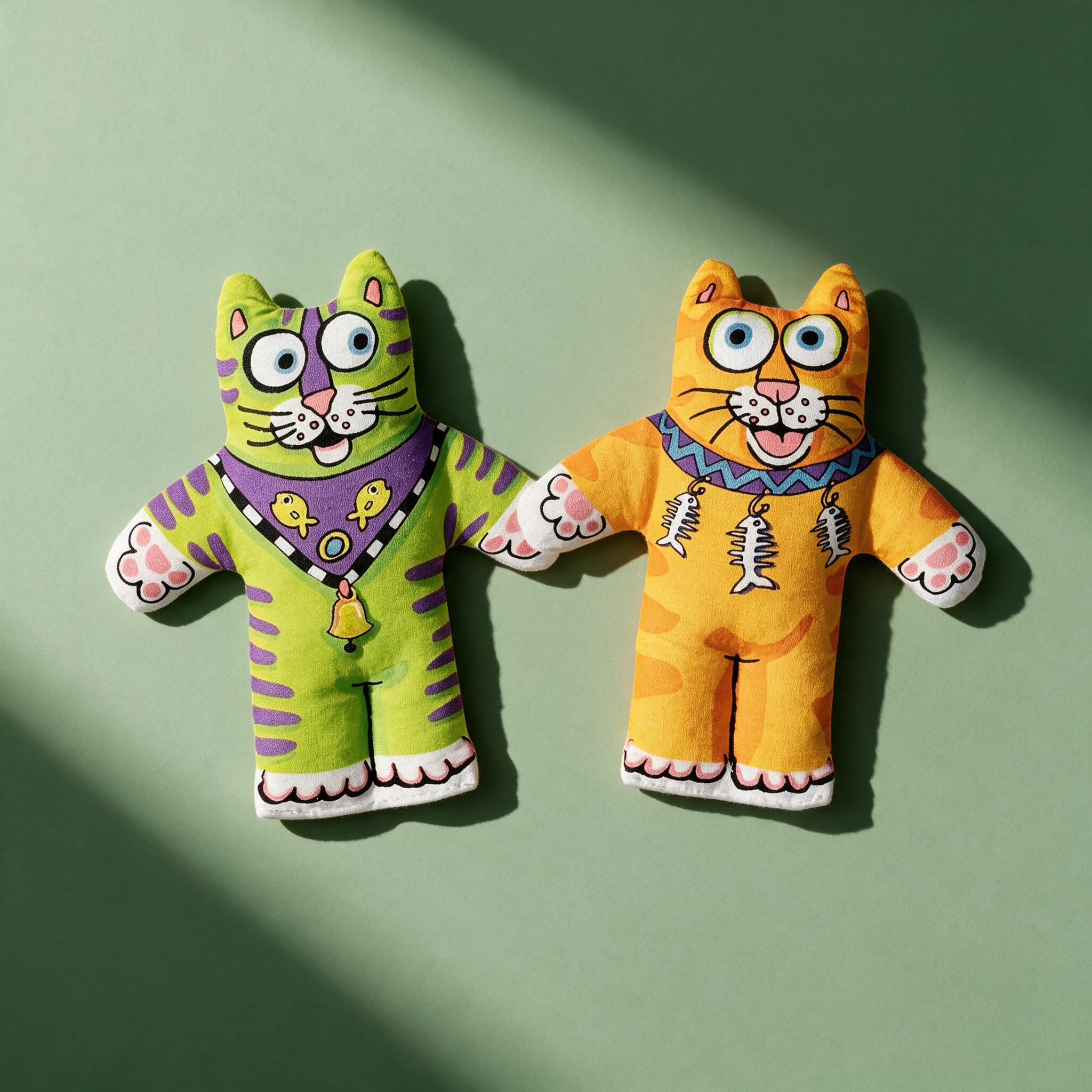Two Fat Cat Kitten toys laid out together on a green background, one in a green tiger print with a purple bandana and bell, the other in an orange tiger print with fish-bone charms.