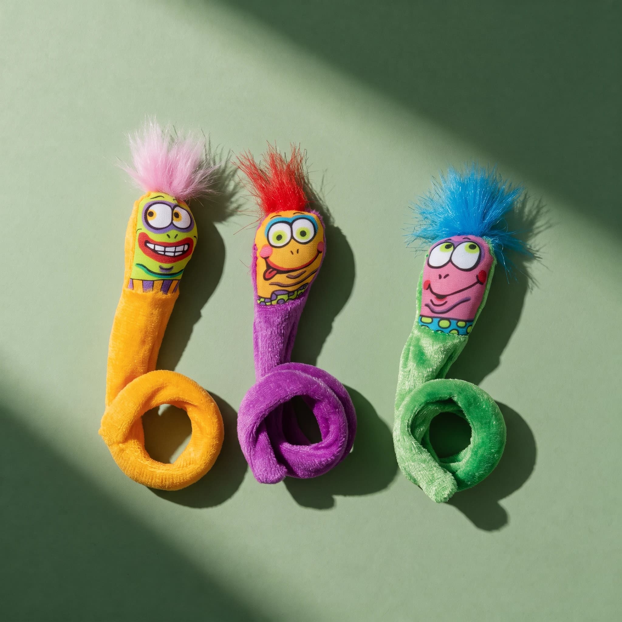 Overhead shot of three Fat Cat Springy Worms cat toys in orange, purple and green, each with a coiled spring tail and a different colourful cartoon face and fuzzy hair tuft, arranged on a sage green background.