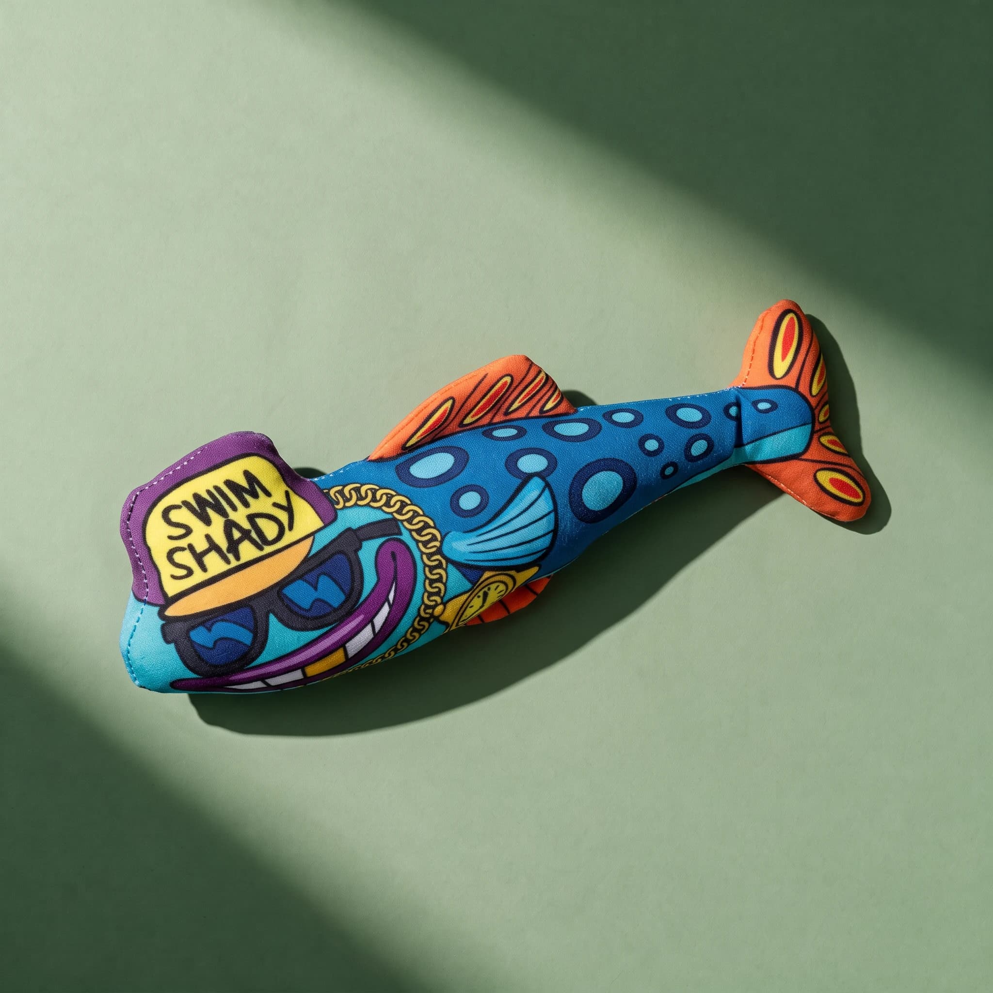 Overhead shot of the Fat Cat Swim Shady plush fish toy, printed with a cartoon rapper-style face in yellow cap, purple sunglasses and gold chain, blue scaled body and orange fins, on a sage green background.
