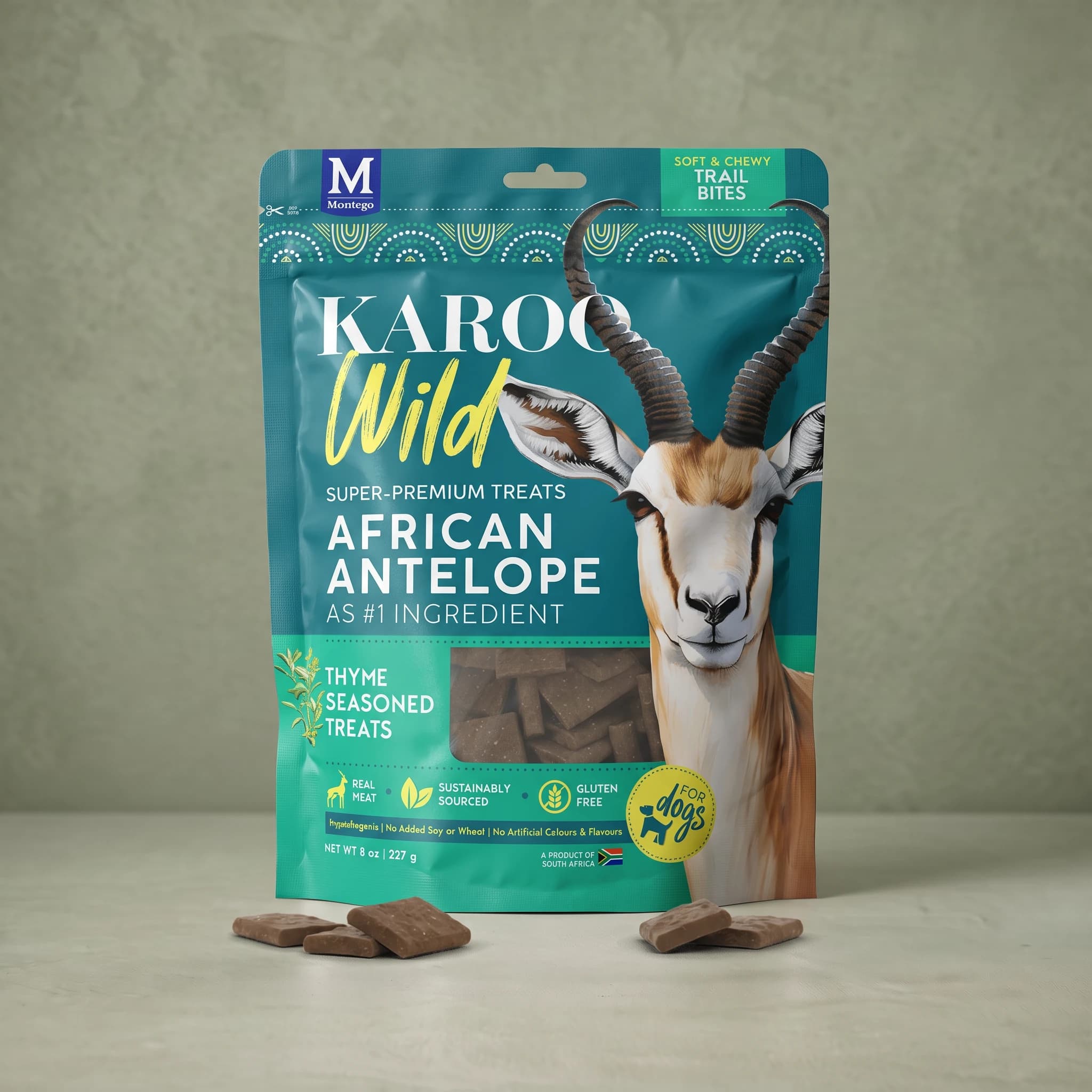 The same teal and green Karoo Wild Thyme Trail Bites pouch standing upright against a plain sage green background, with small square dark brown treats scattered at its base.