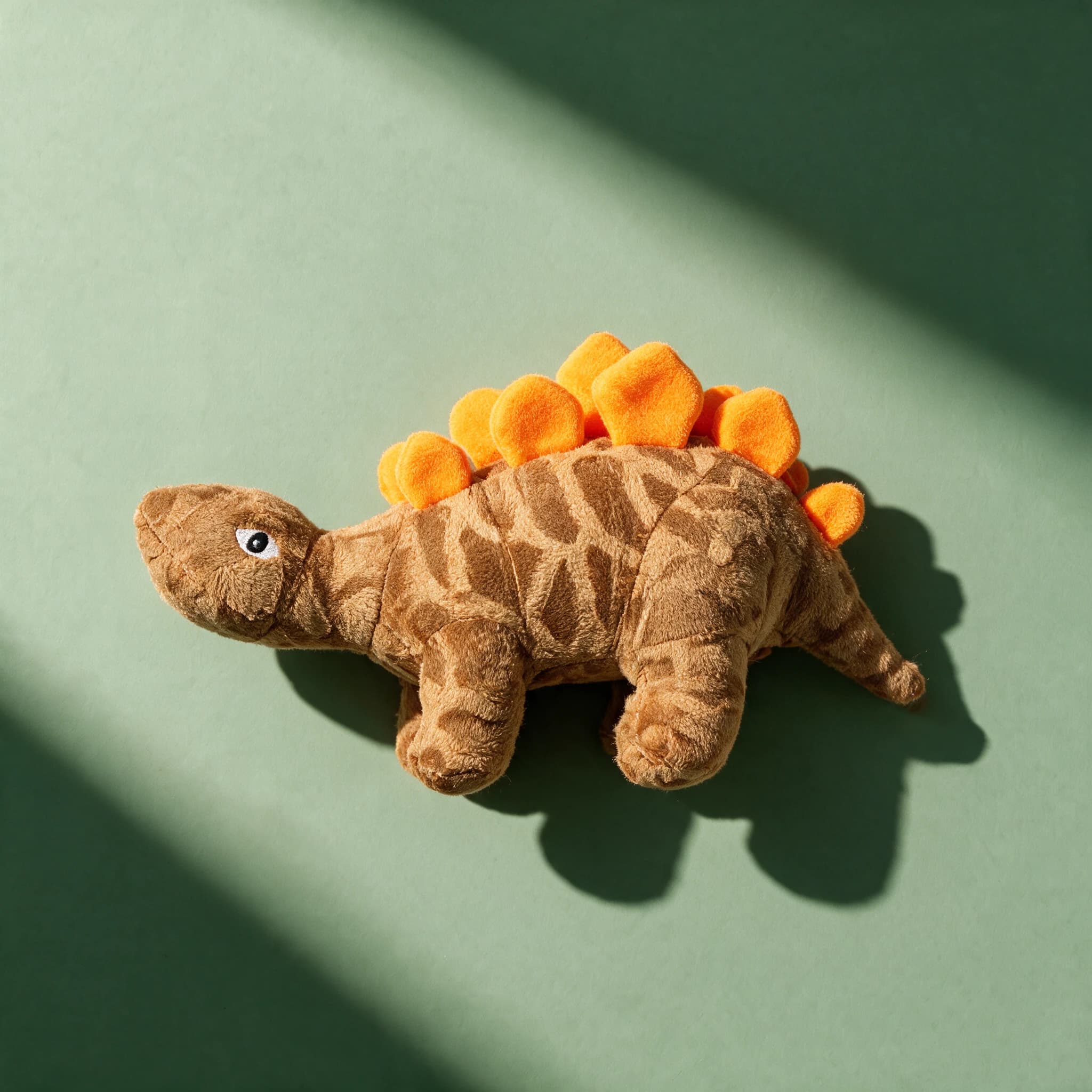 Overhead shot of the brown patterned plush Mighty Toys Dinosaur Stego with orange spiked back, positioned beside a grey plush Triceratops-style dinosaur toy with white horns, on a sage green background.