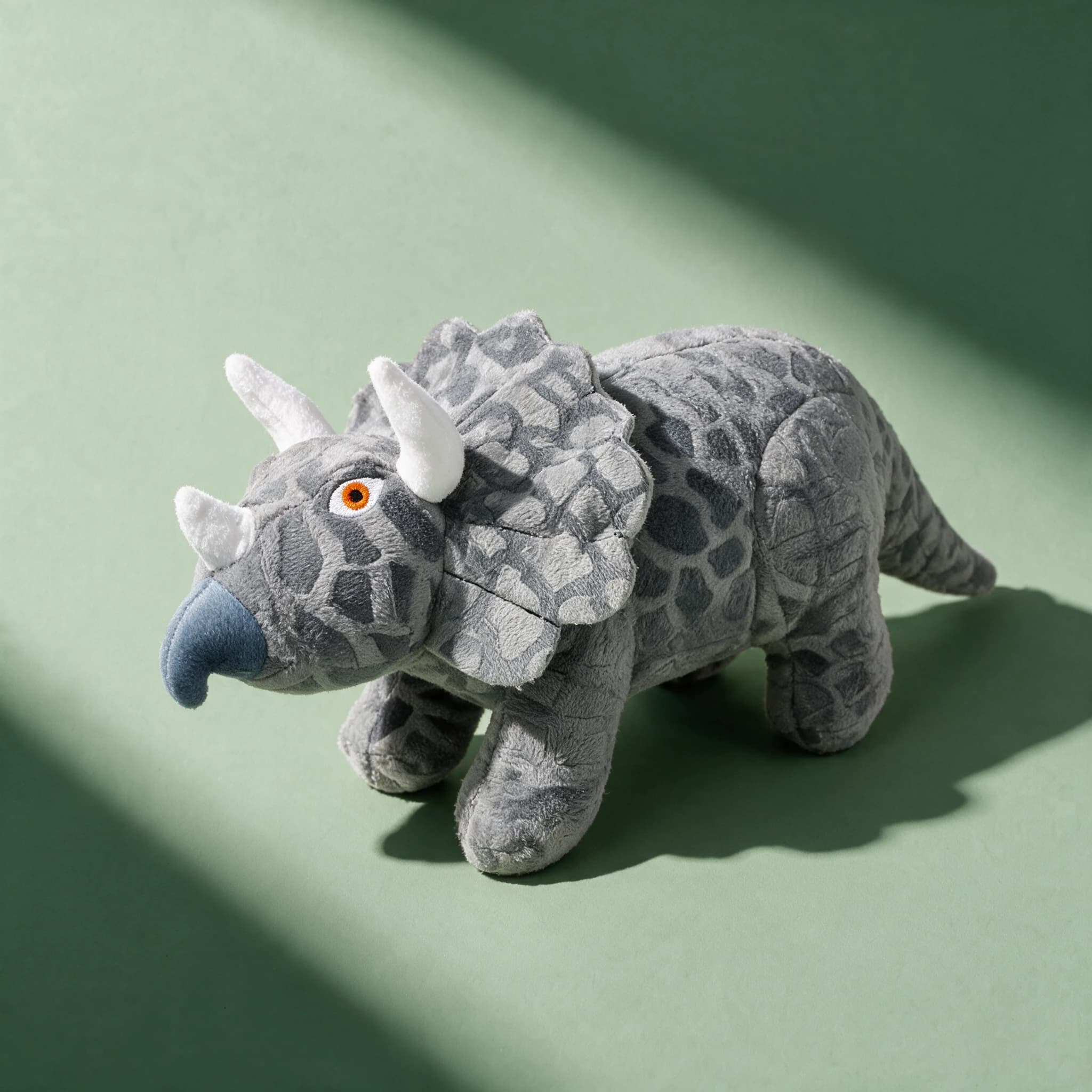 Overhead view of the grey plush Mighty Toys Dinosaur Tri triceratops toy, featuring a textured grey mottled body, white horns, a blue beak, and an embroidered orange eye.