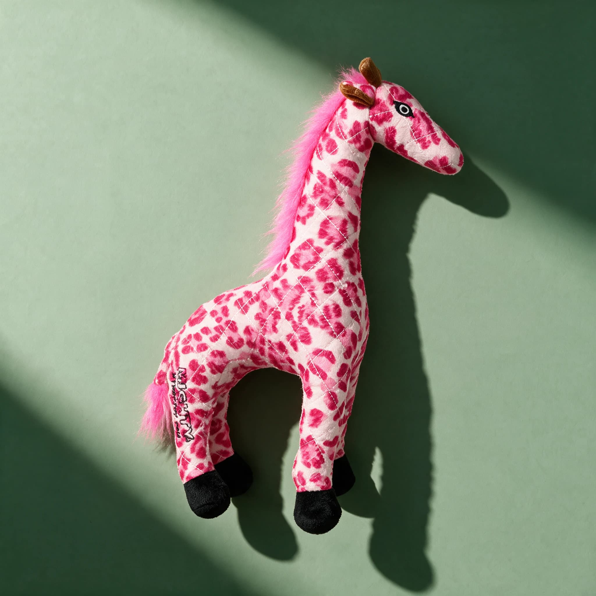 The pink leopard-print Mighty Toys Safari Giraffe plush toy, with a long quilted neck, black hooves, brown horns, and a pink fuzzy mane and tail, against a sage green background.