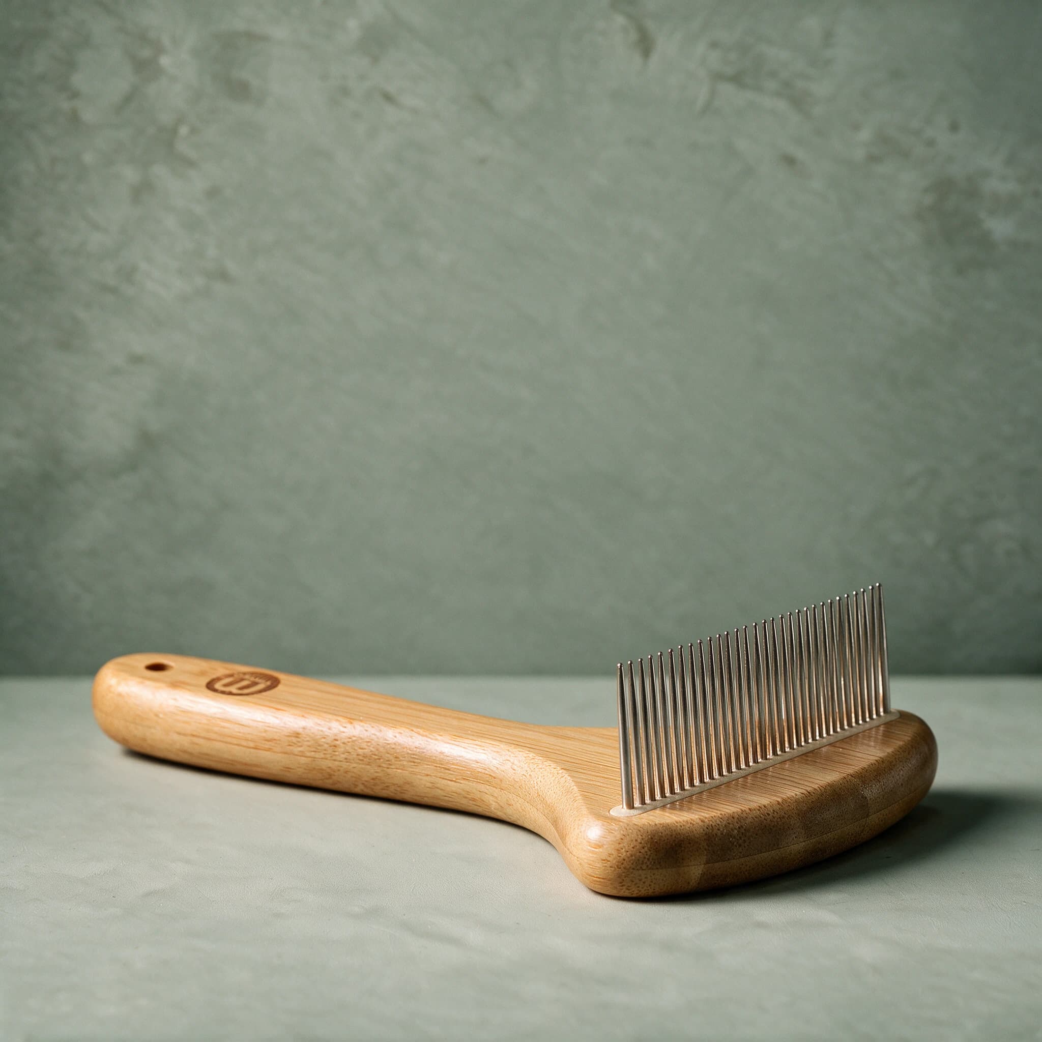 Mikki Bamboo Anti-Tangle Comb with a bamboo handle and a row of long, rounded stainless steel teeth, shown on a sage green background.
