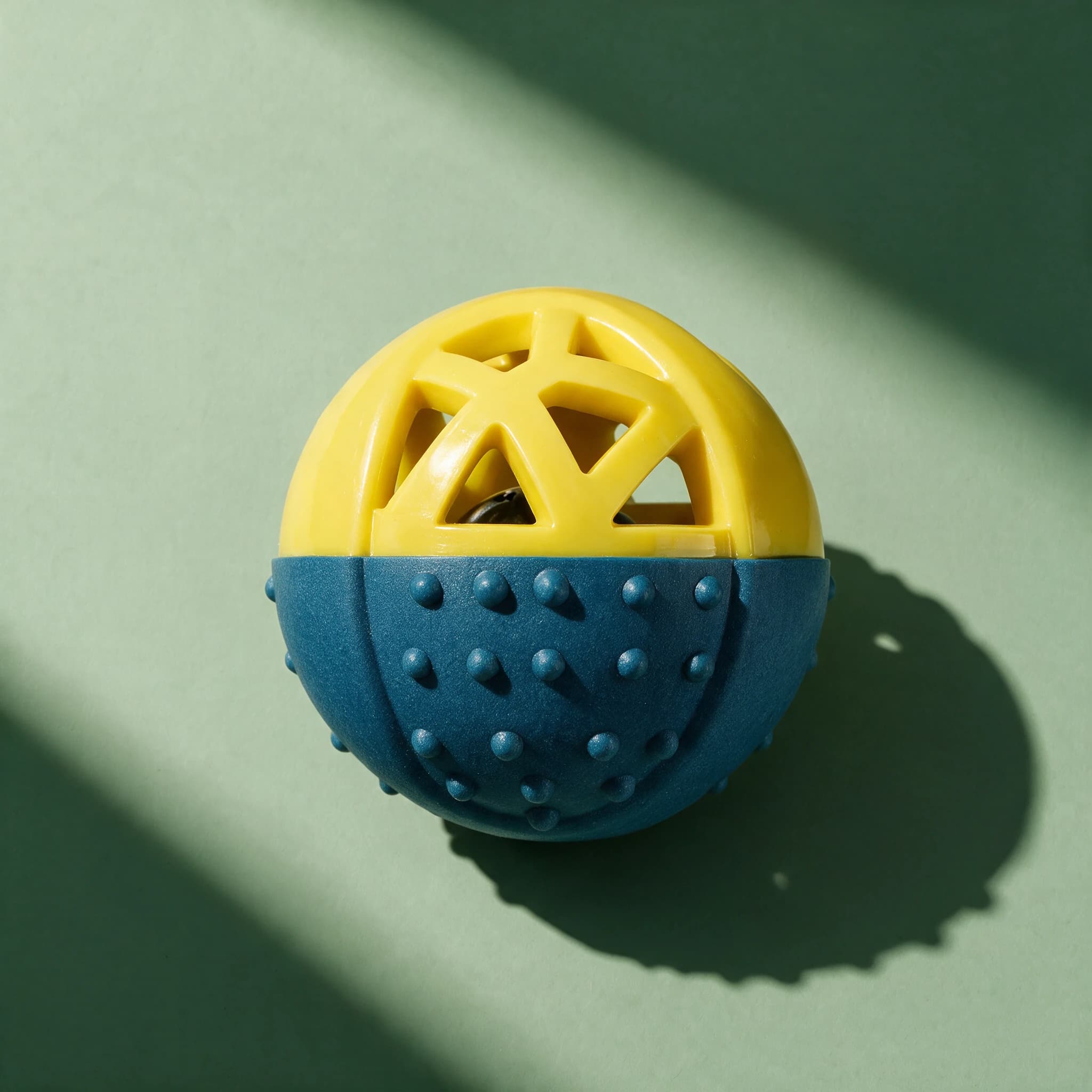 Close-up of the yellow and blue PetLove Nuzzle Puzzle Ball with its openwork geometric top and dotted rubber base, shown alone against a green background.