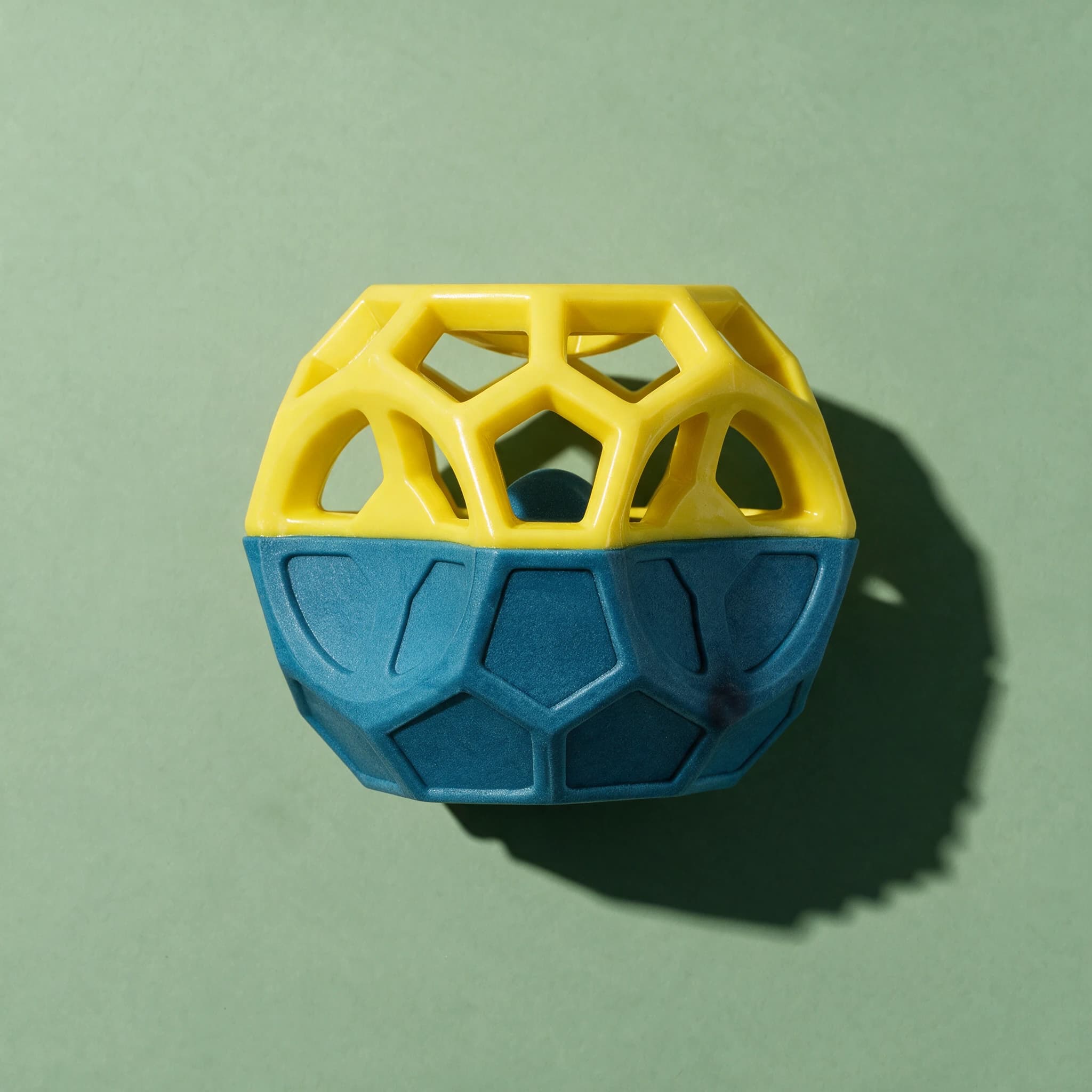 Overhead view of the yellow and teal PetLove Nuzzle Puzzle Cube, showing its faceted lattice top and hexagon-textured base, on a sage-green background with a soft shadow.