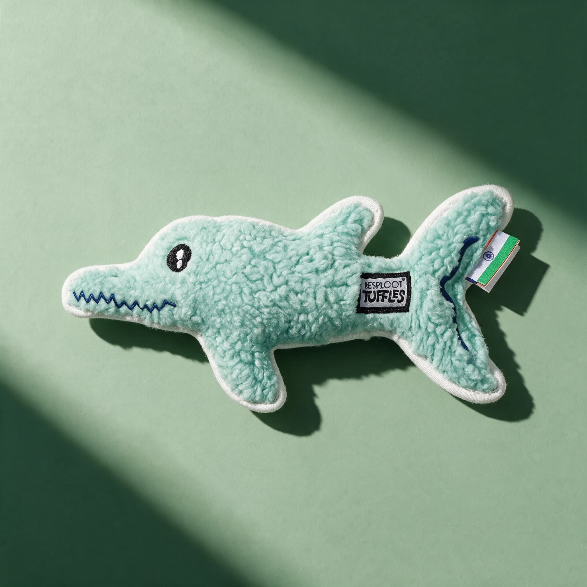 Close-up of the mint green fleece Tuffles dolphin toy alone, showing its fuzzy sherpa texture, stitched black eye, zigzag mouth, white edge piping and Resploot Tuffles fabric label, on a sage green background.