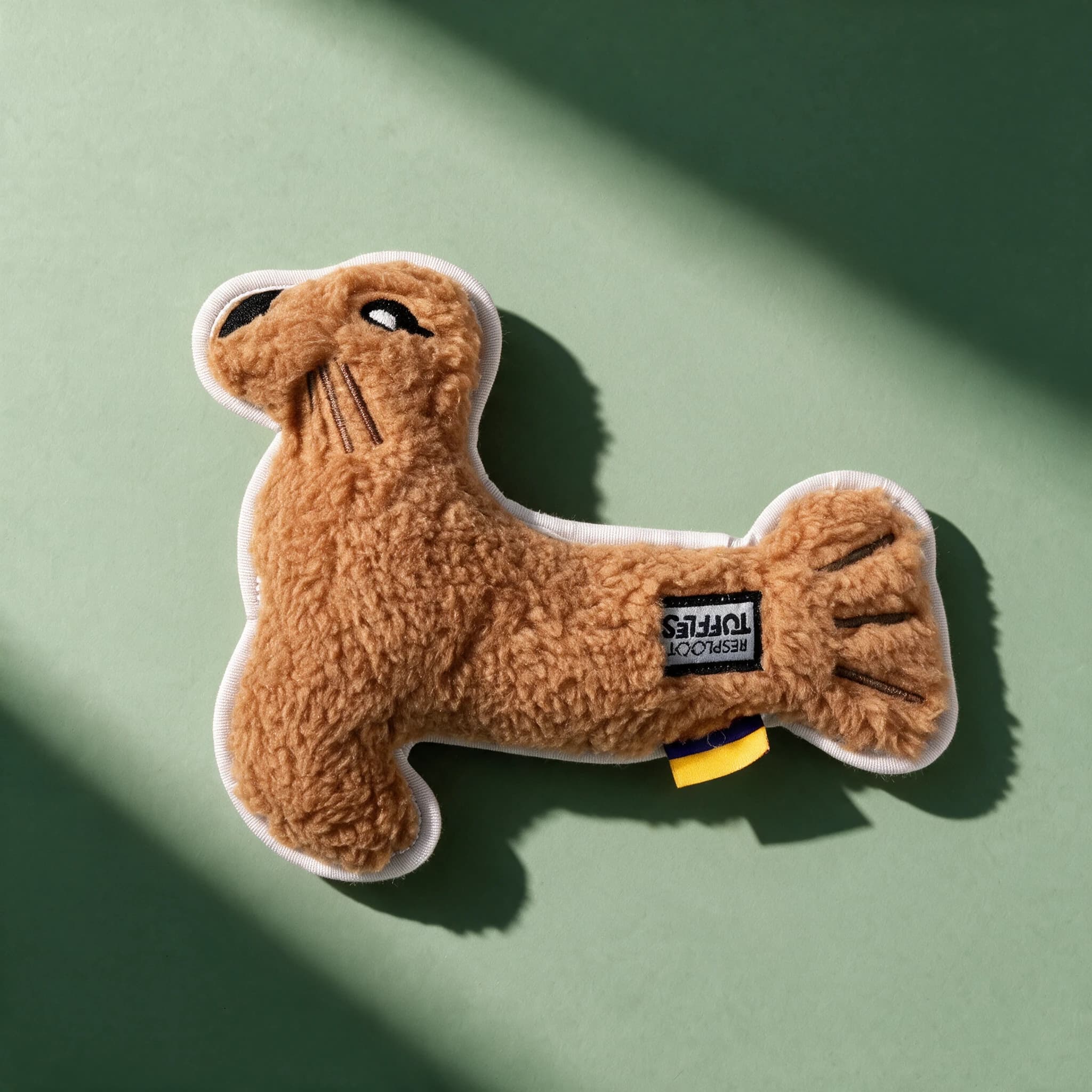 Overhead view of the tan fleece PetLove Resploot Tuffles Sea Lion toy, showing its fuzzy textured body, embroidered whiskers, stitched claws, and a yellow and navy fabric tag.