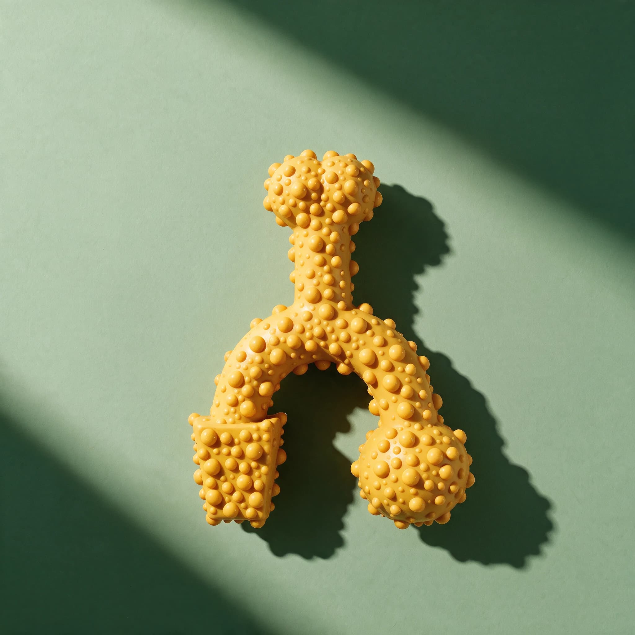 Close-up overhead view of the mustard-yellow Tasty Bone Braai Chicken toy, showing its dense bumpy, studded surface texture, on a sage-green background with a strong diagonal shadow.