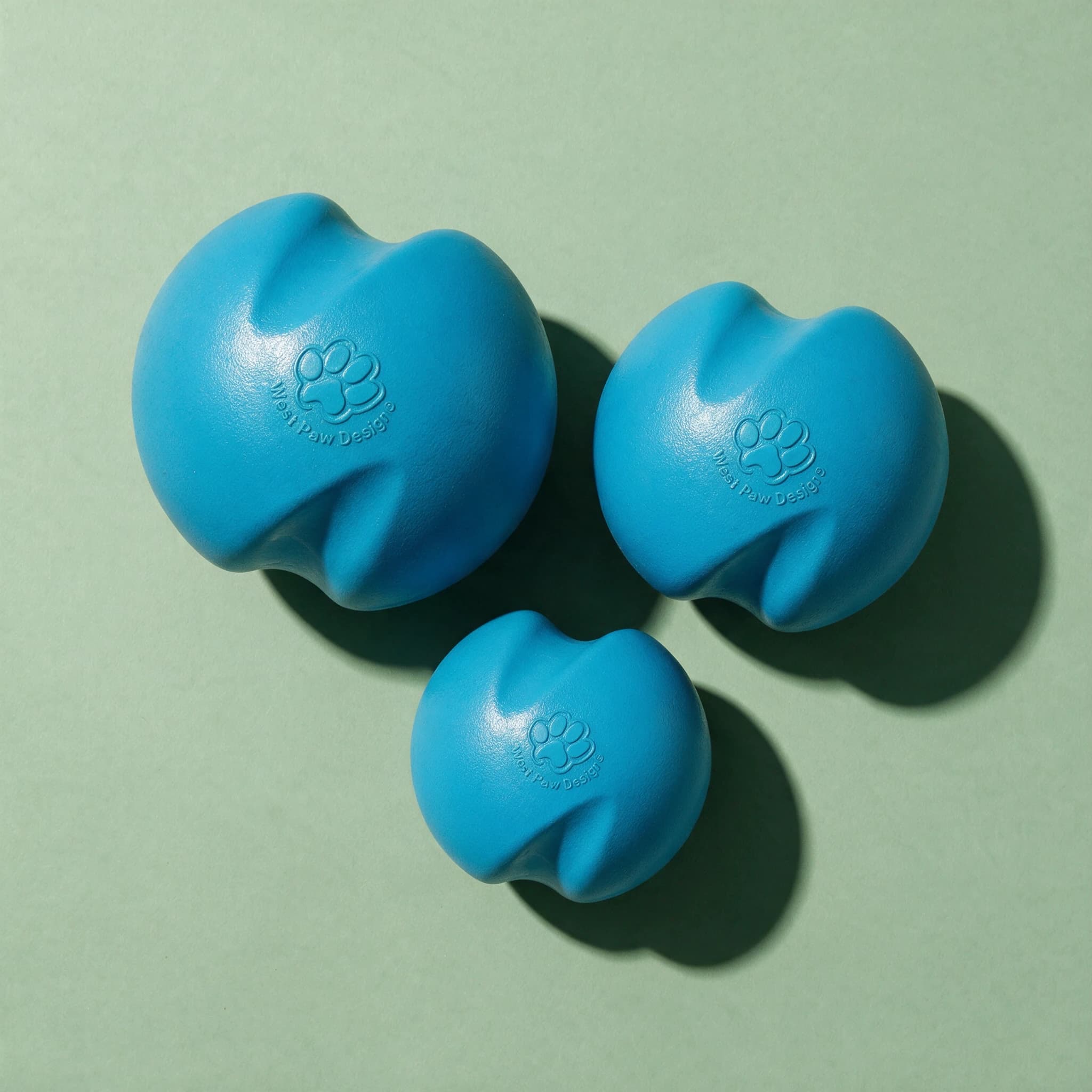 Overhead view of three West Paw Jive Dog Balls in different sizes, all in matching teal blue with a wavy organic shape and embossed West Paw Design paw print logo, arranged together against a sage green background with strong diagonal shadows.