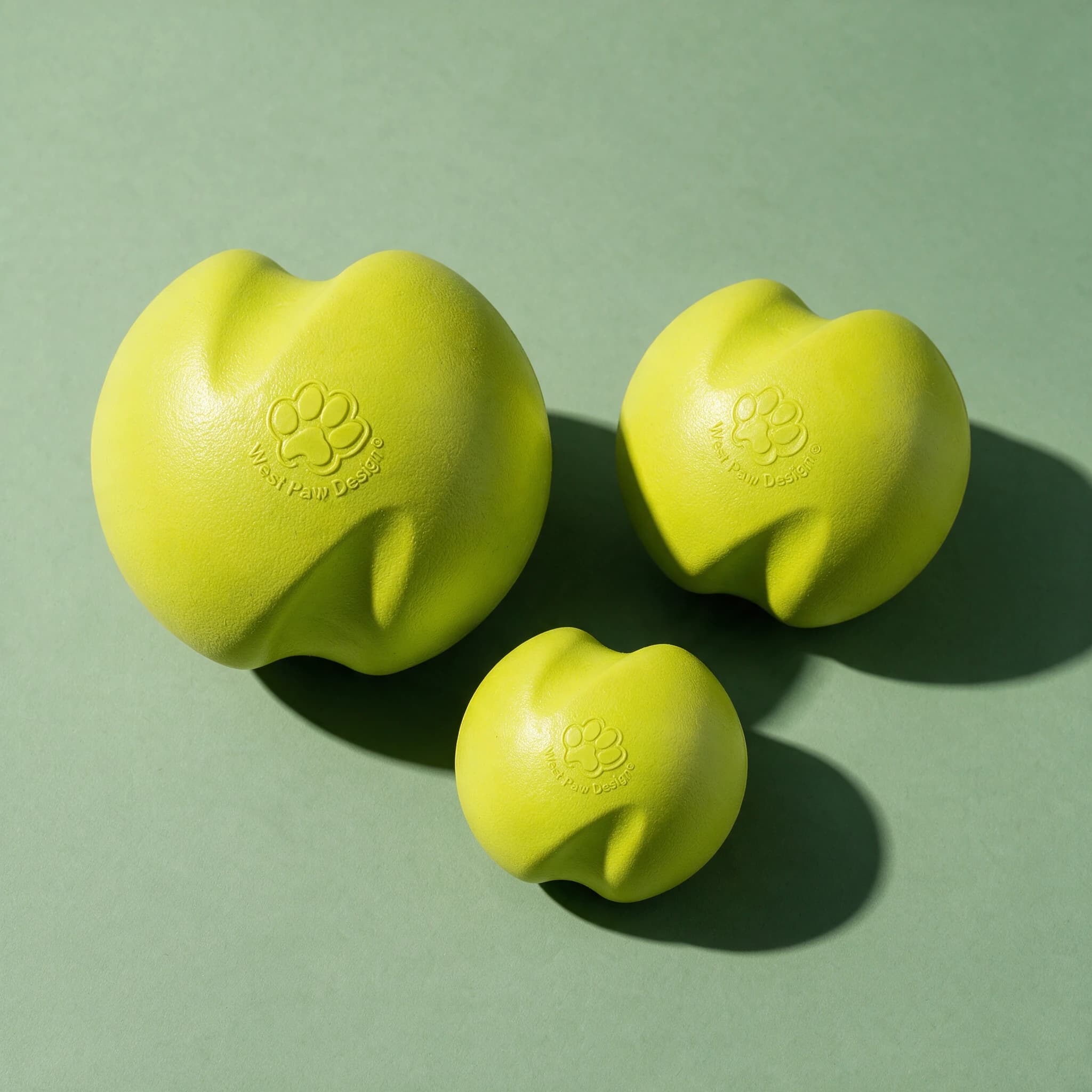 Overhead view of three West Paw Jive Dog Balls in different sizes, all in matching lime green with a wavy organic shape and embossed West Paw Design paw print logo, arranged together against a sage green background with strong diagonal shadows.