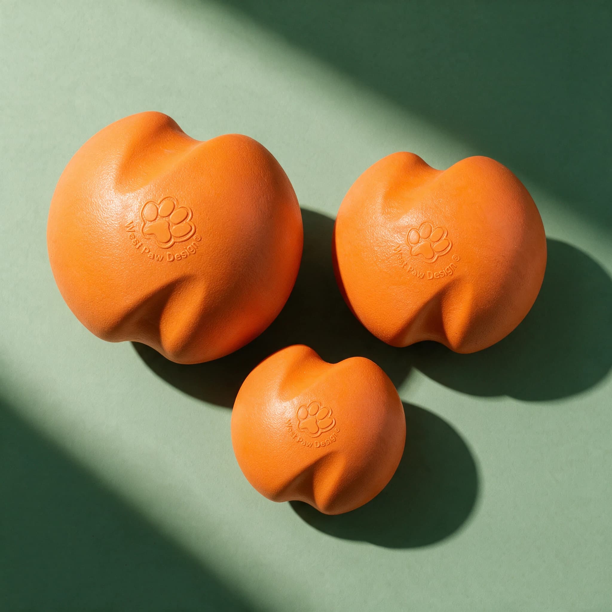 Overhead view of three West Paw Jive Dog Balls in different sizes, all in matching orange with a wavy organic shape and embossed West Paw Design paw print logo, arranged together against a sage green background with strong diagonal shadows.