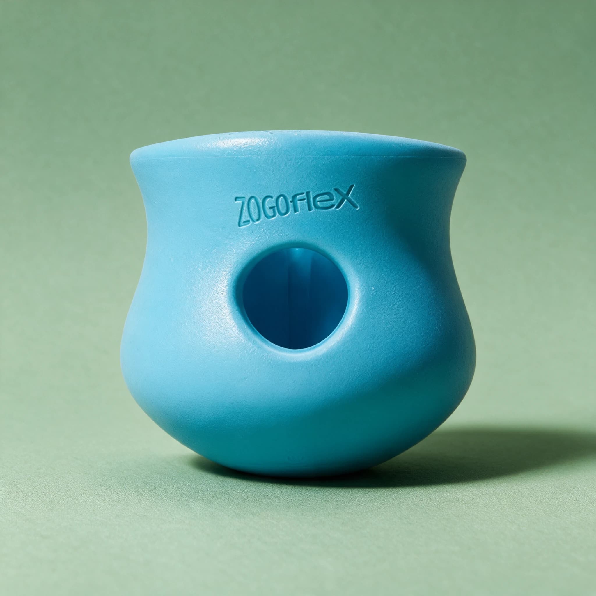 A blue West Paw Toppl treat toy, a curved cup-shaped rubber toy embossed with the Zogoflex logo and featuring a round hole through its side, photographed against a sage-green background.