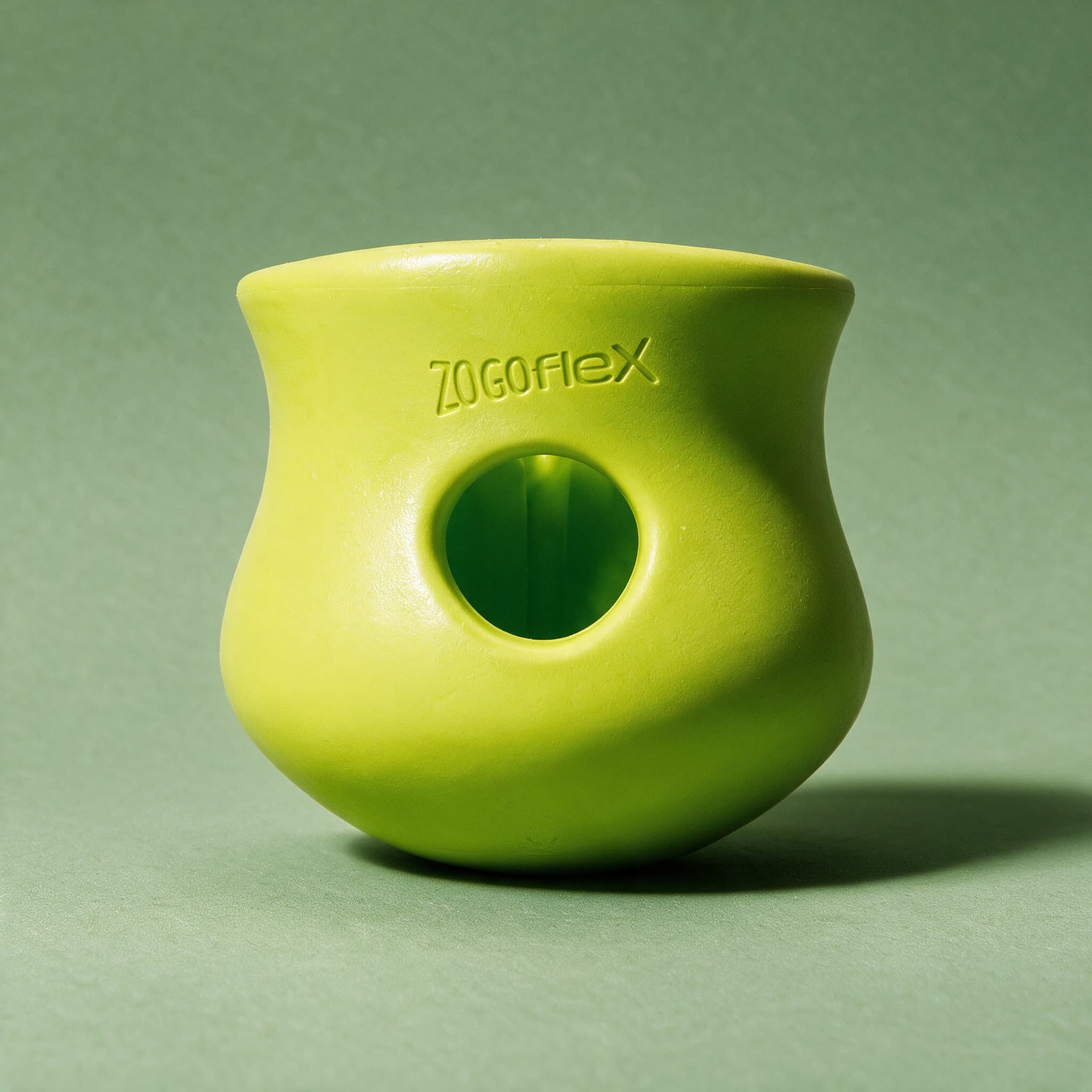 Close-up of a single green West Paw Zogoflex Toppl treat toy, showing its curved vase-like shape, round side opening and embossed Zogoflex logo, against a sage green background.